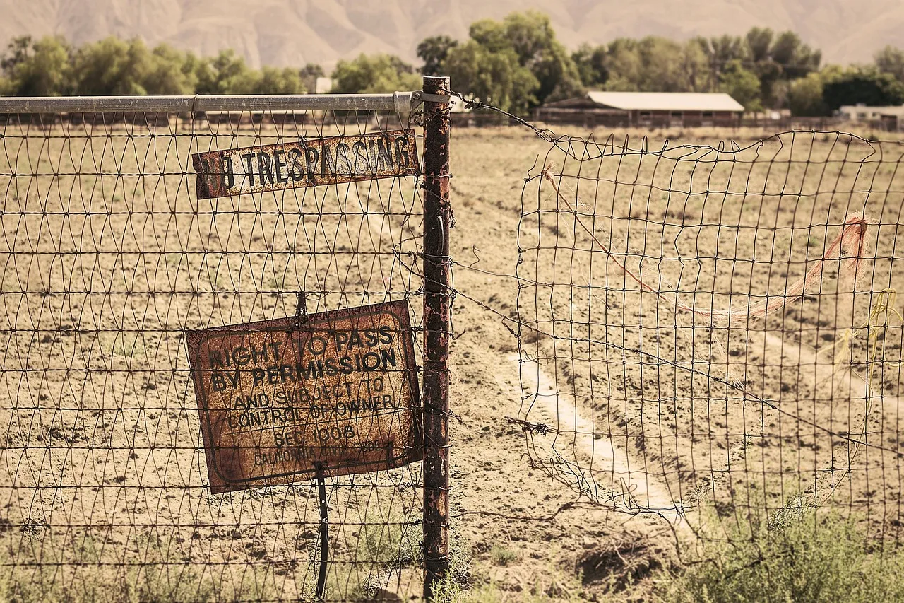A weathered NO TRESPASSING sign and a second sign reading RIGHT TO PASS BY PERMISSION, both nailed to a post on a sagging wire fence. To the right of the post the fence has been pulled open, leaving a gap into the dry field beyond.