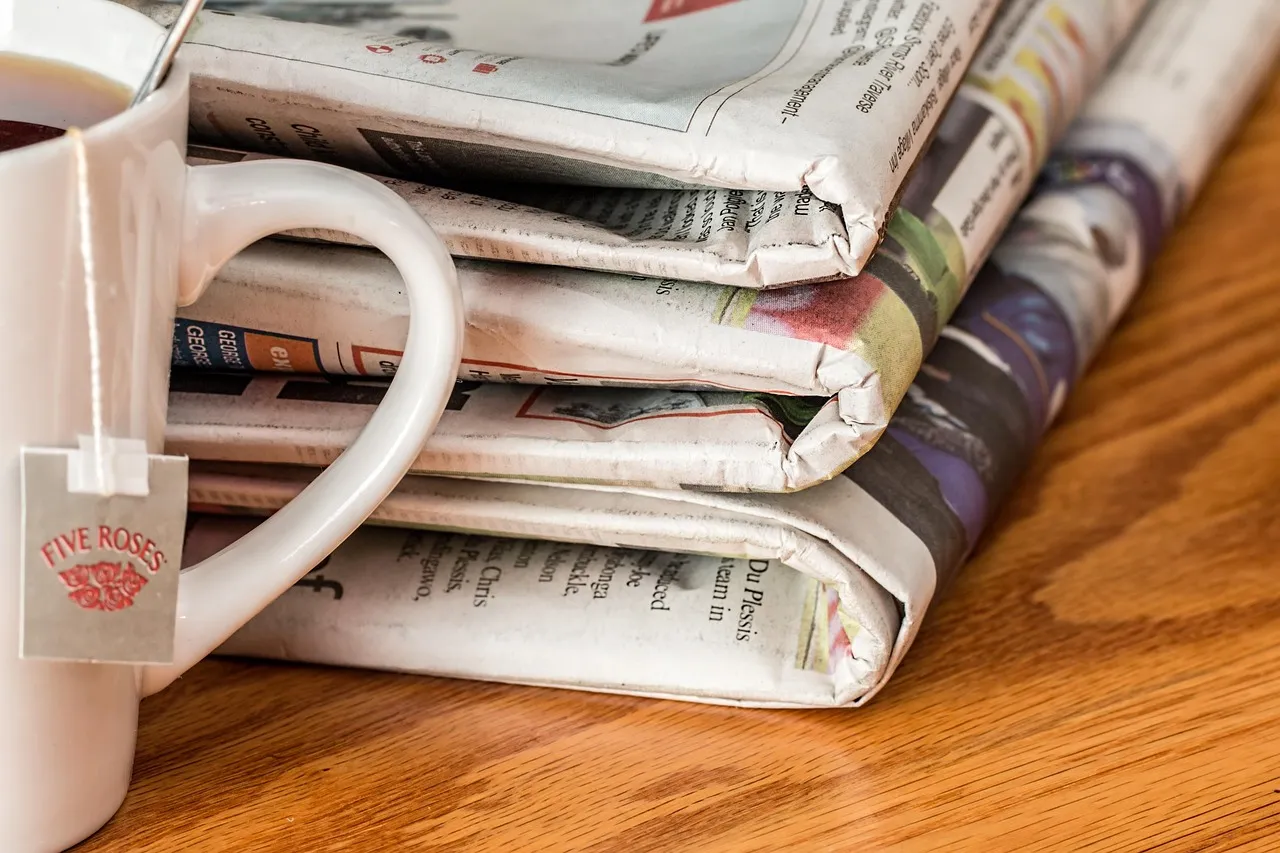 Illustrative: a stack of folded printed newspapers on a wooden table beside a mug.