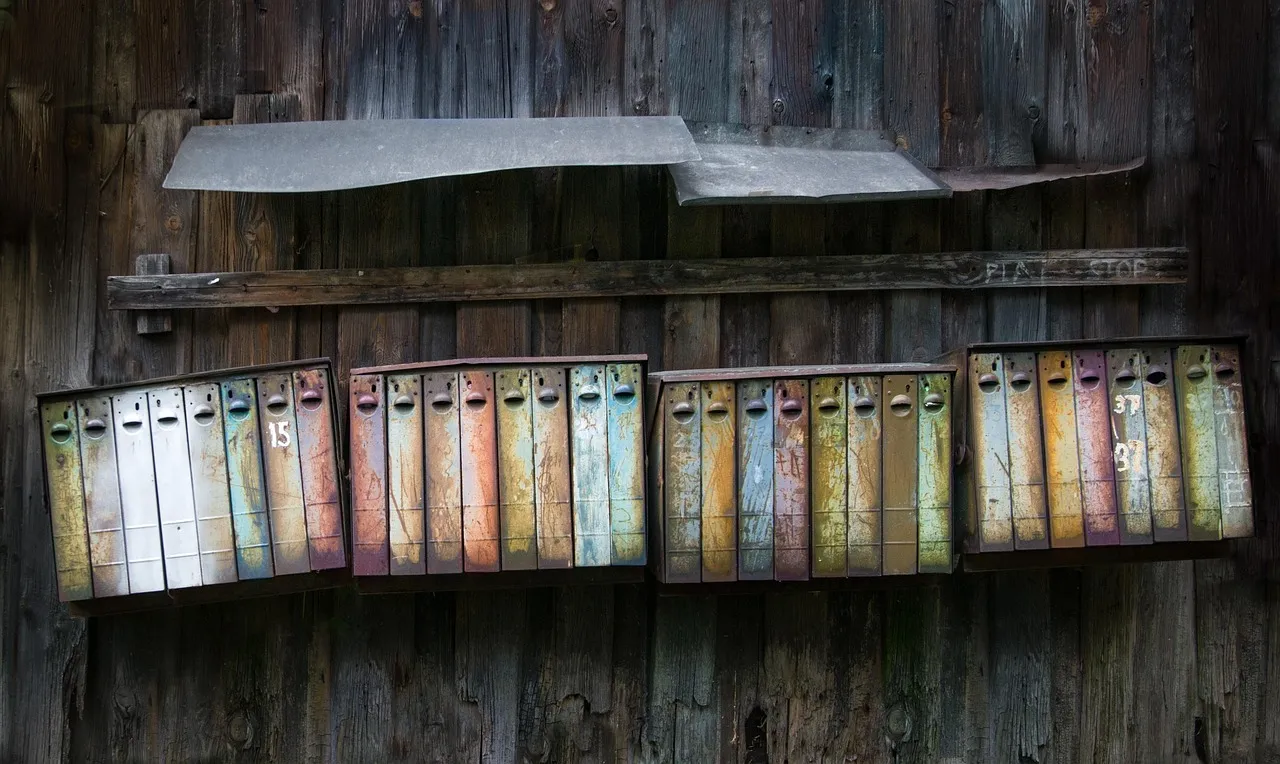 Four banks of narrow metal mailboxes mounted on a weathered wooden wall, each box a different faded colour and several numbered by hand, with a rusted tin canopy above them.