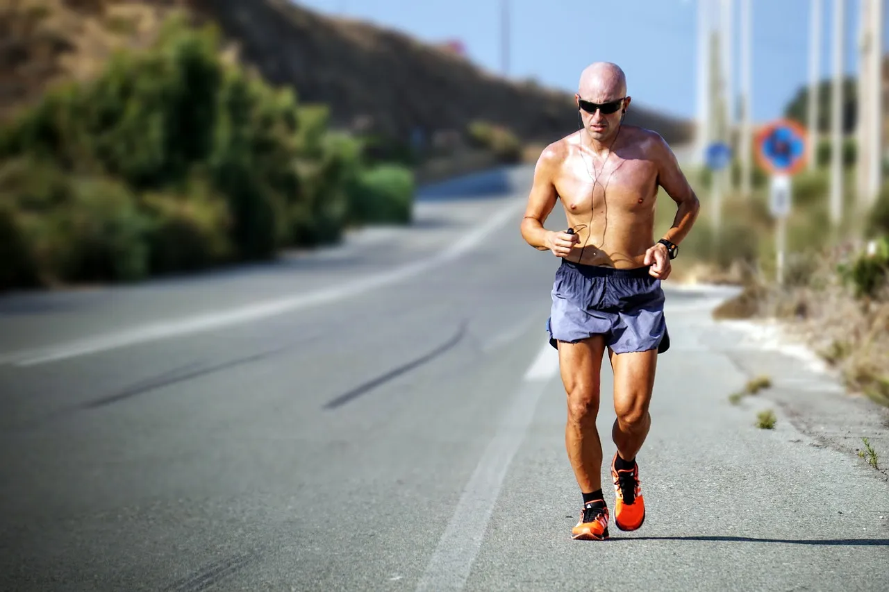 A runner on a sunlit road in the heat, shirt off, mid-stride