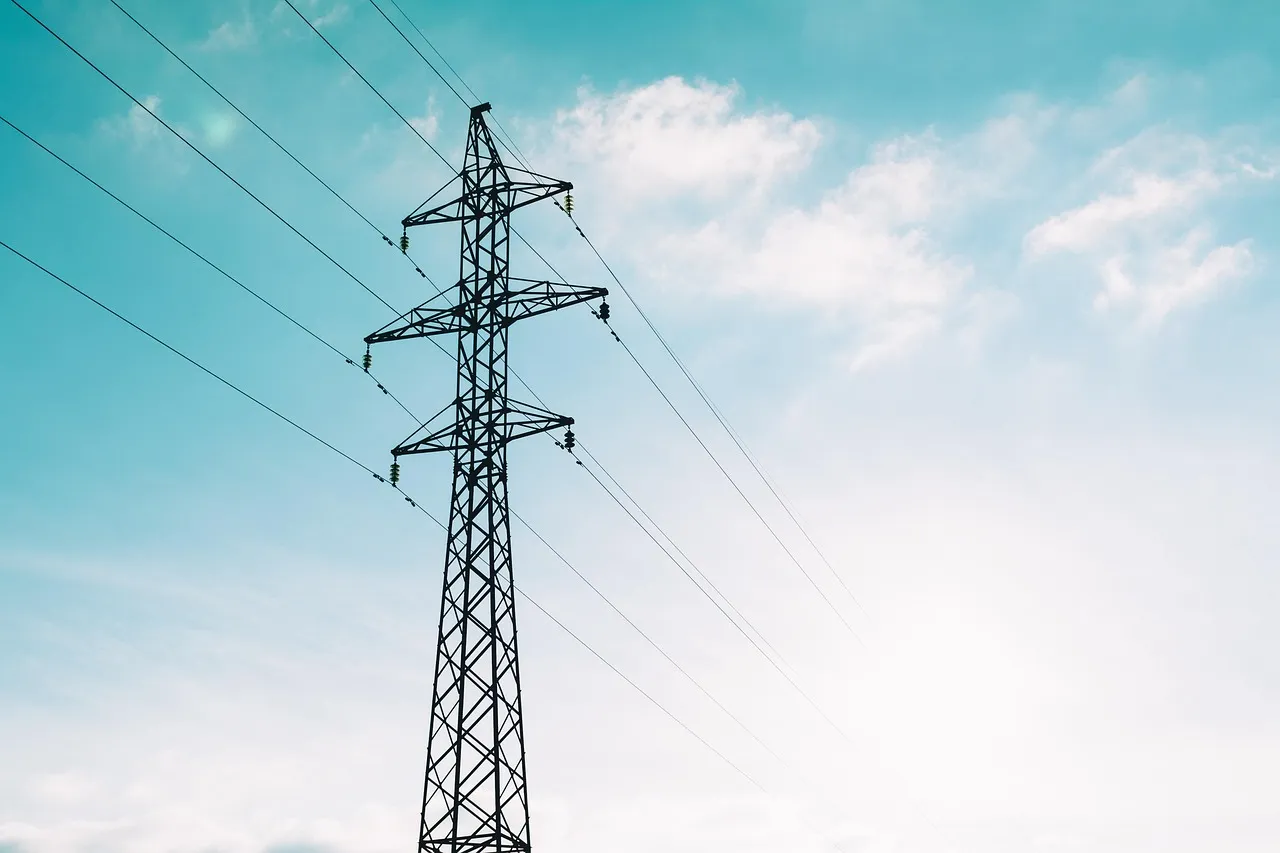 A single steel transmission pylon carrying several conductor lines, silhouetted against a pale blue sky with light cloud