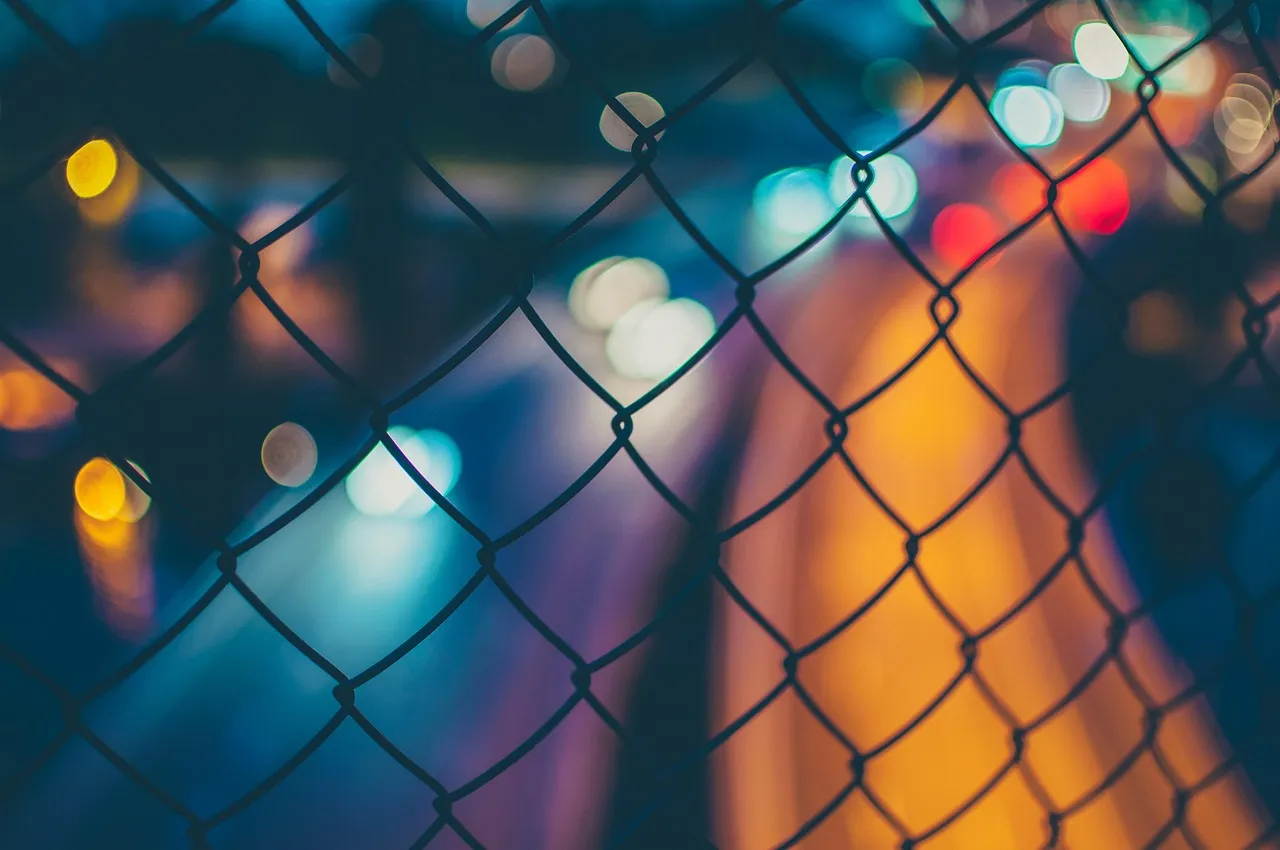 A chain-link fence in sharp focus with city lights blurred beyond it
