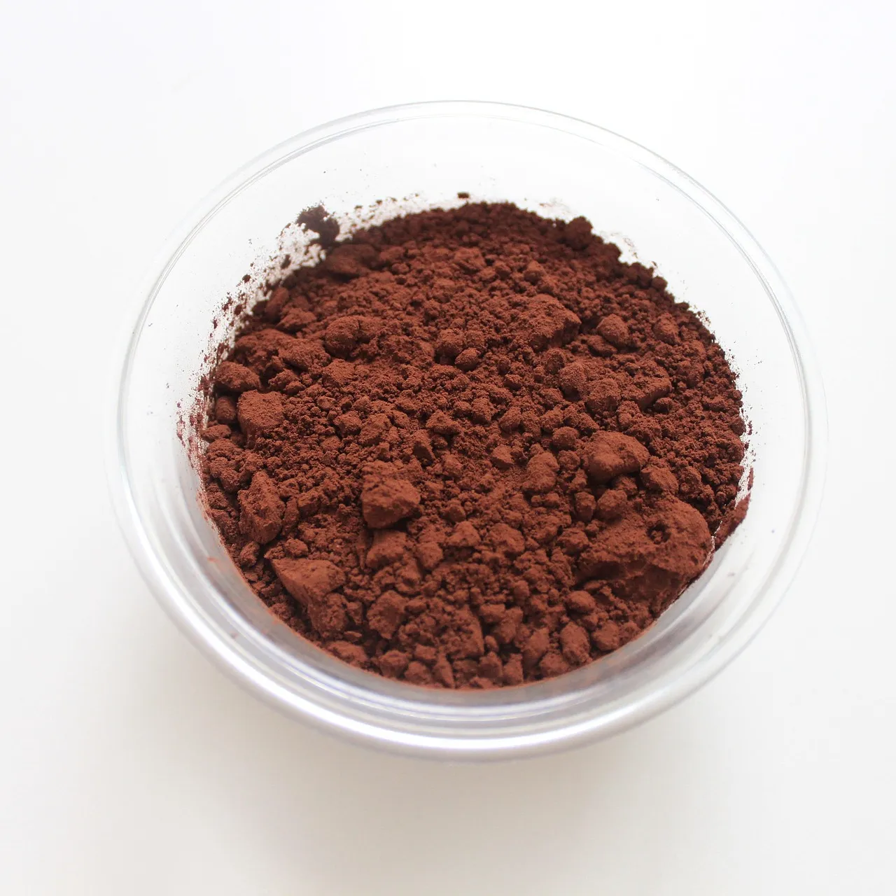 A small glass bowl of loose reddish-brown cocoa powder photographed from above on a plain white surface.