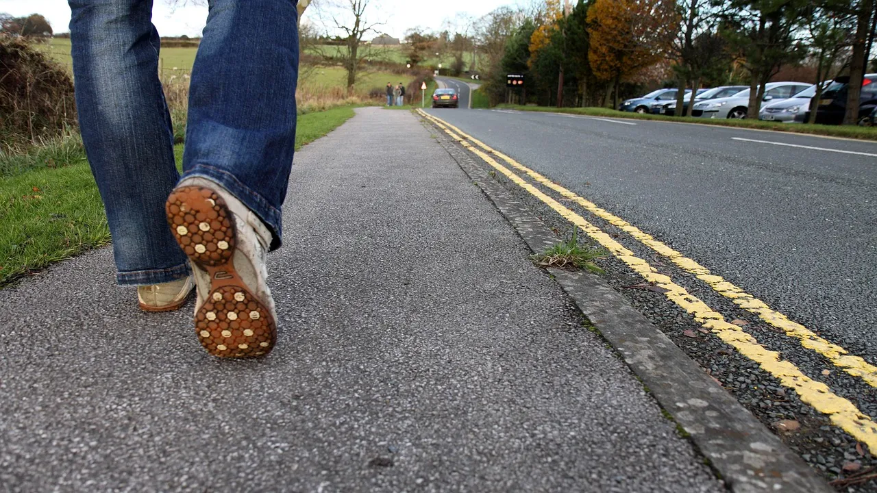 A low-angle view of someone walking along the edge of a country road, one boot mid-stride, double yellow lines running into the distance.