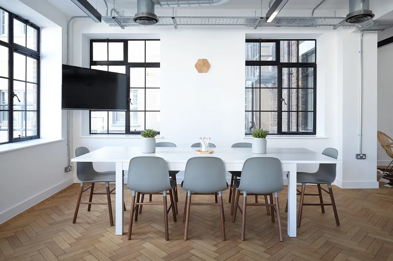 An empty meeting room with a long white table, grey chairs and a wall-mounted screen, lit by two windows