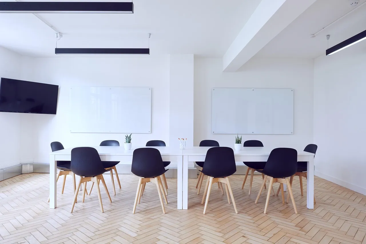 An empty meeting room with a long white table, moulded chairs and a blank wall-mounted screen.