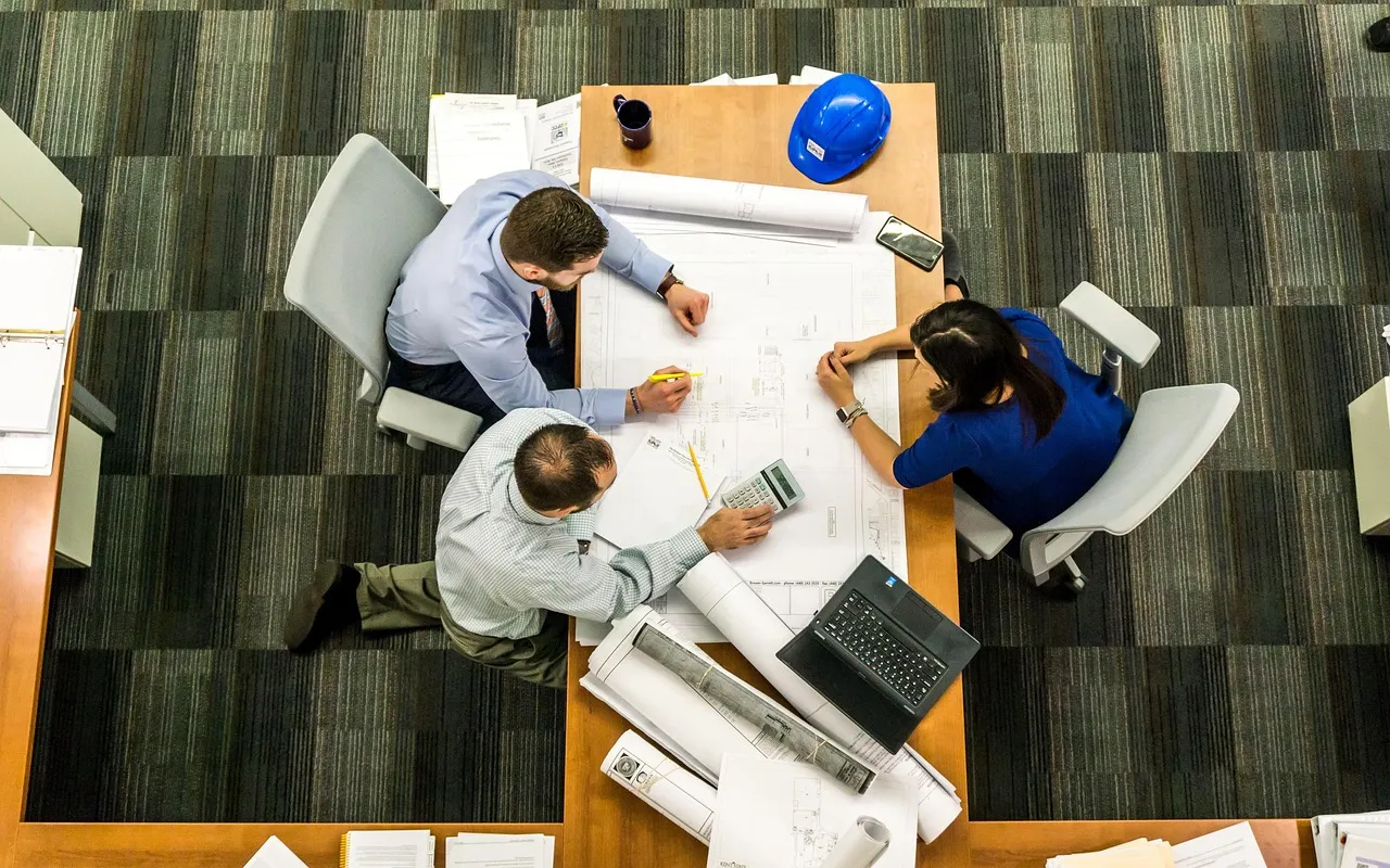 Three people reviewing technical drawings around a table, seen from above