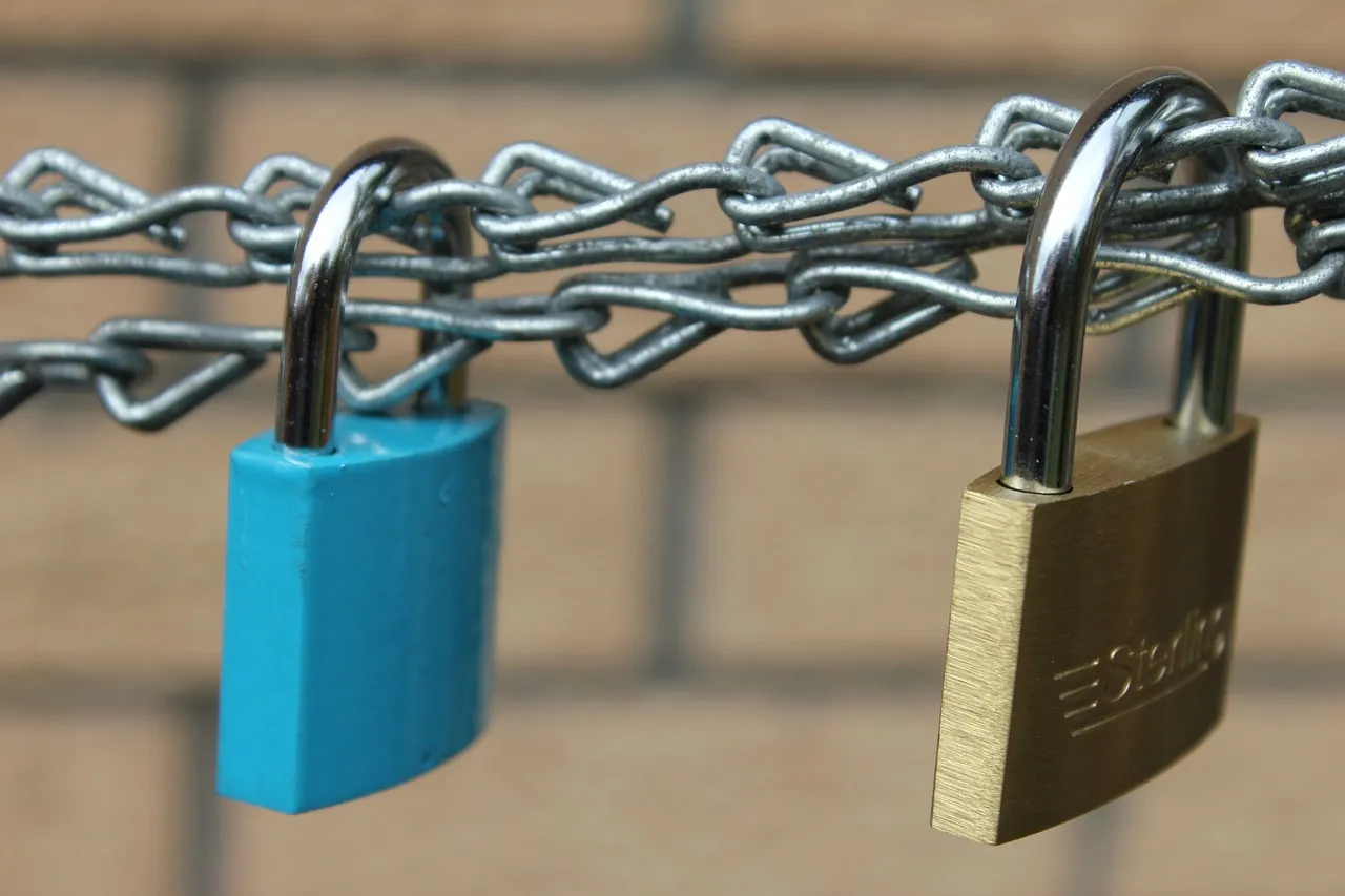 Two padlocks, one blue and one brass, hanging from a chain against a brick wall.
