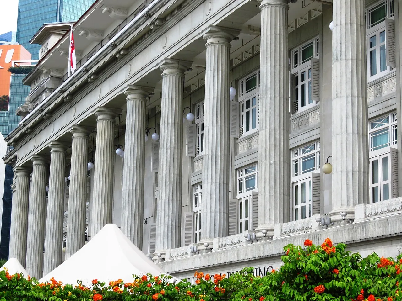 The colonnaded stone facade of a classical Singapore building, a Singapore flag flying at the left and flowering shrubs below