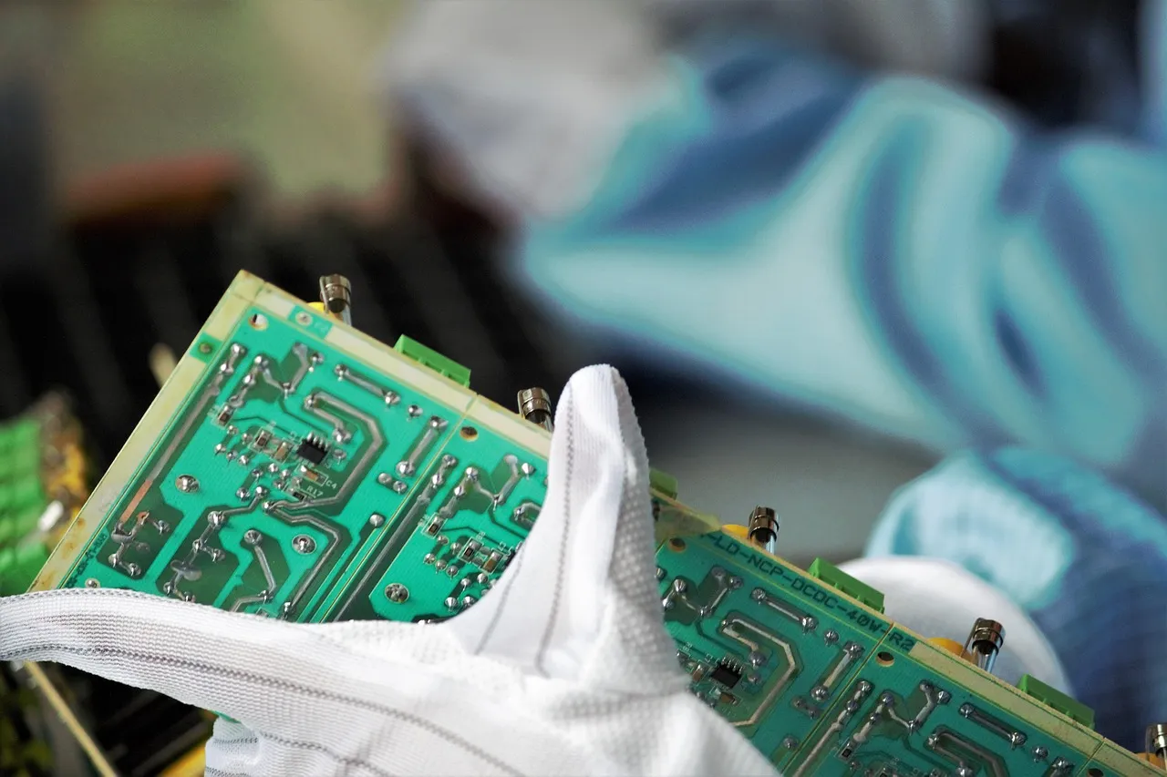 Gloved hands holding a green circuit board on an electronics production line, illustrating a report on high-bandwidth memory pricing and supply.