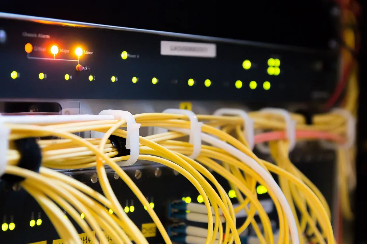 A rack-mounted network chassis with its alarm panel lit, critical and major indicators glowing amber, and bundled yellow fibre patch cables running across the front