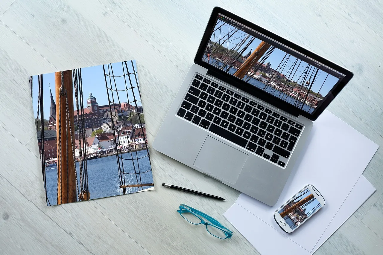 A desk from above with a laptop, a phone and a printed photograph all showing the same harbour image