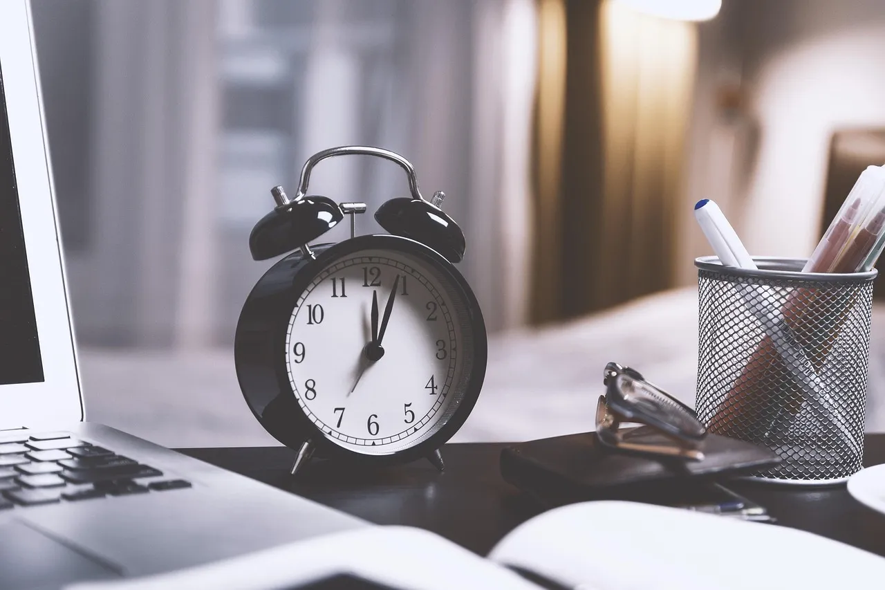 A black alarm clock on a desk beside a laptop, showing a few minutes to twelve.
