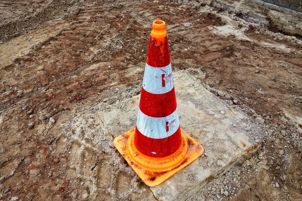 A weathered orange and white traffic cone standing alone on a patch of concrete at a dug-up construction site