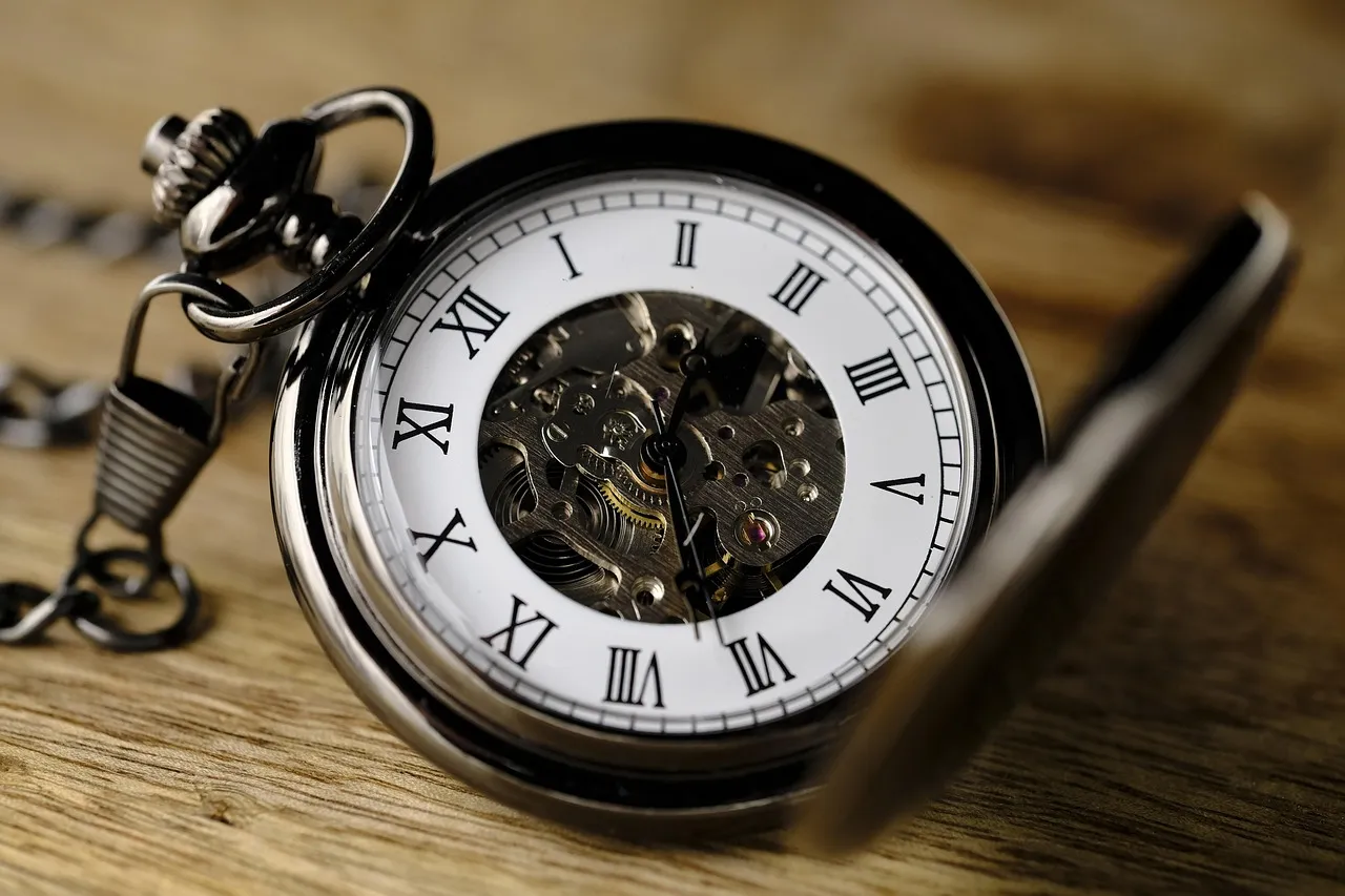 An open pocket watch with roman numerals resting on a wooden surface