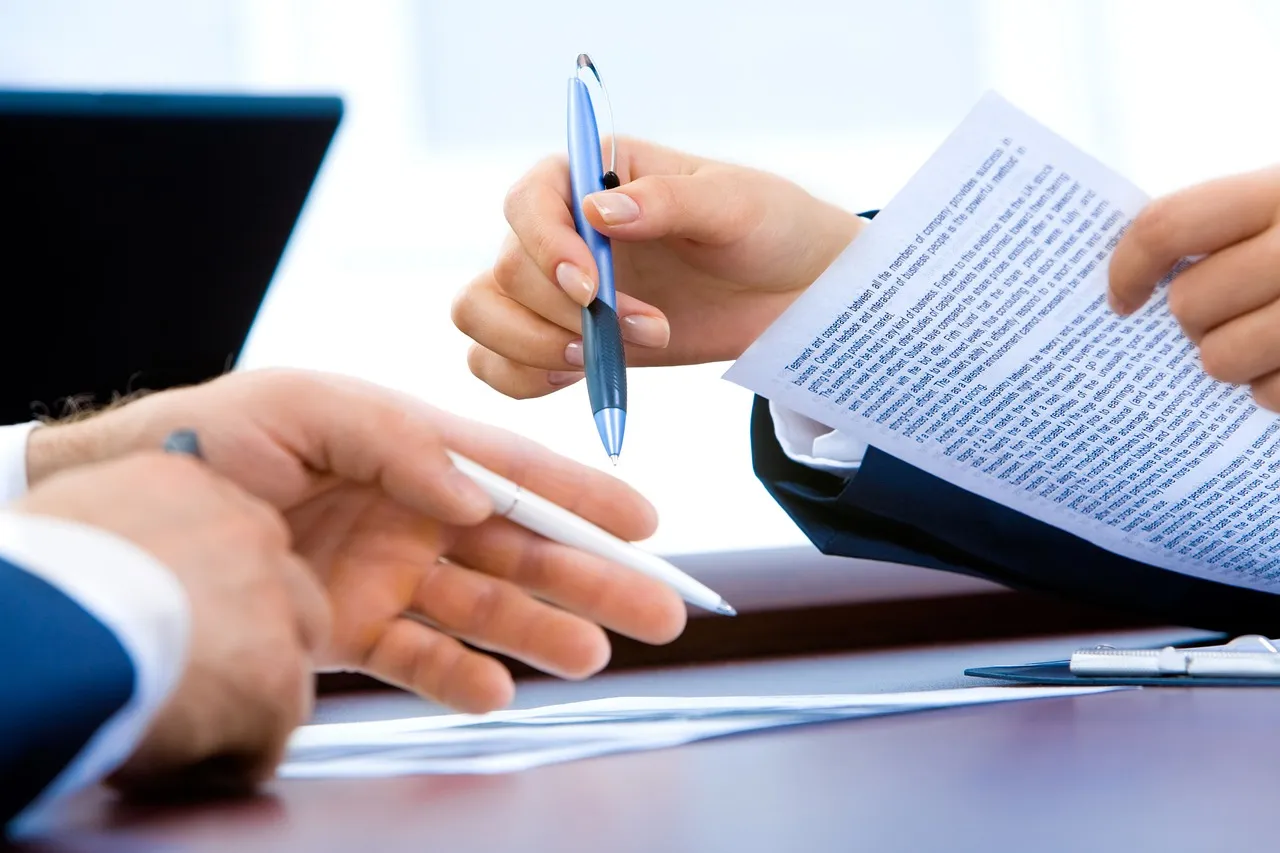 Hands holding a pen over a printed document beside a laptop — illustrating the act of checking a text's origin, which is the part not yet possible.