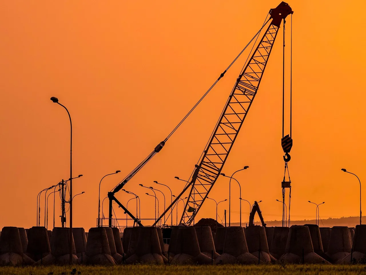 A lattice-boom crawler crane silhouetted against an orange sunset sky above a line of concrete breakwater blocks and street lamps