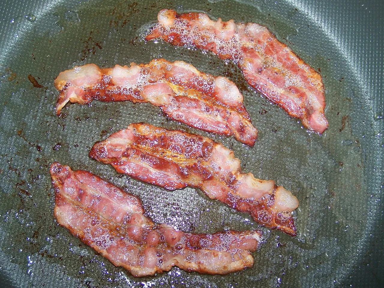 Rashers of bacon frying in a dark non-stick pan.