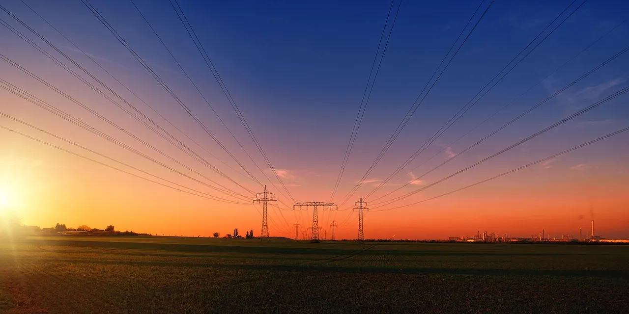 Overhead transmission lines fanning across flat open farmland at sunset, with three lattice pylons on the horizon and an industrial plant beyond them