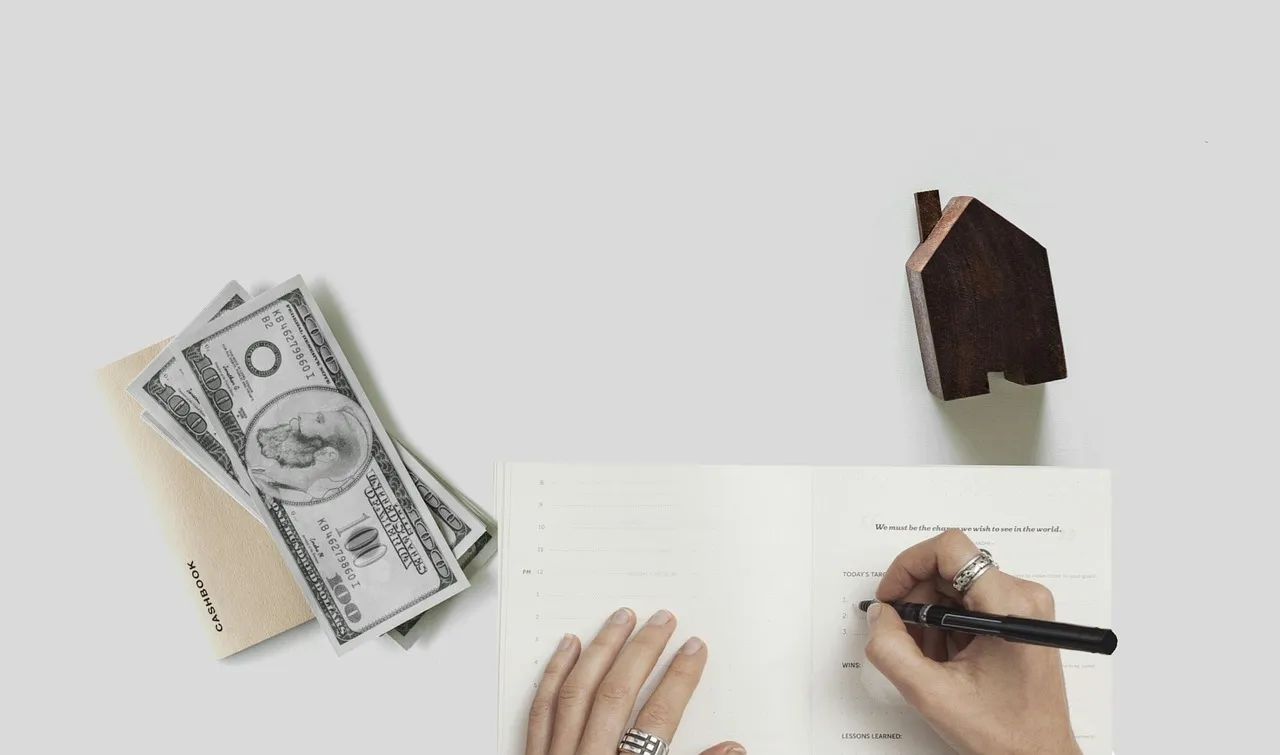 Banknotes and a small wooden house model beside a hand writing on a document.