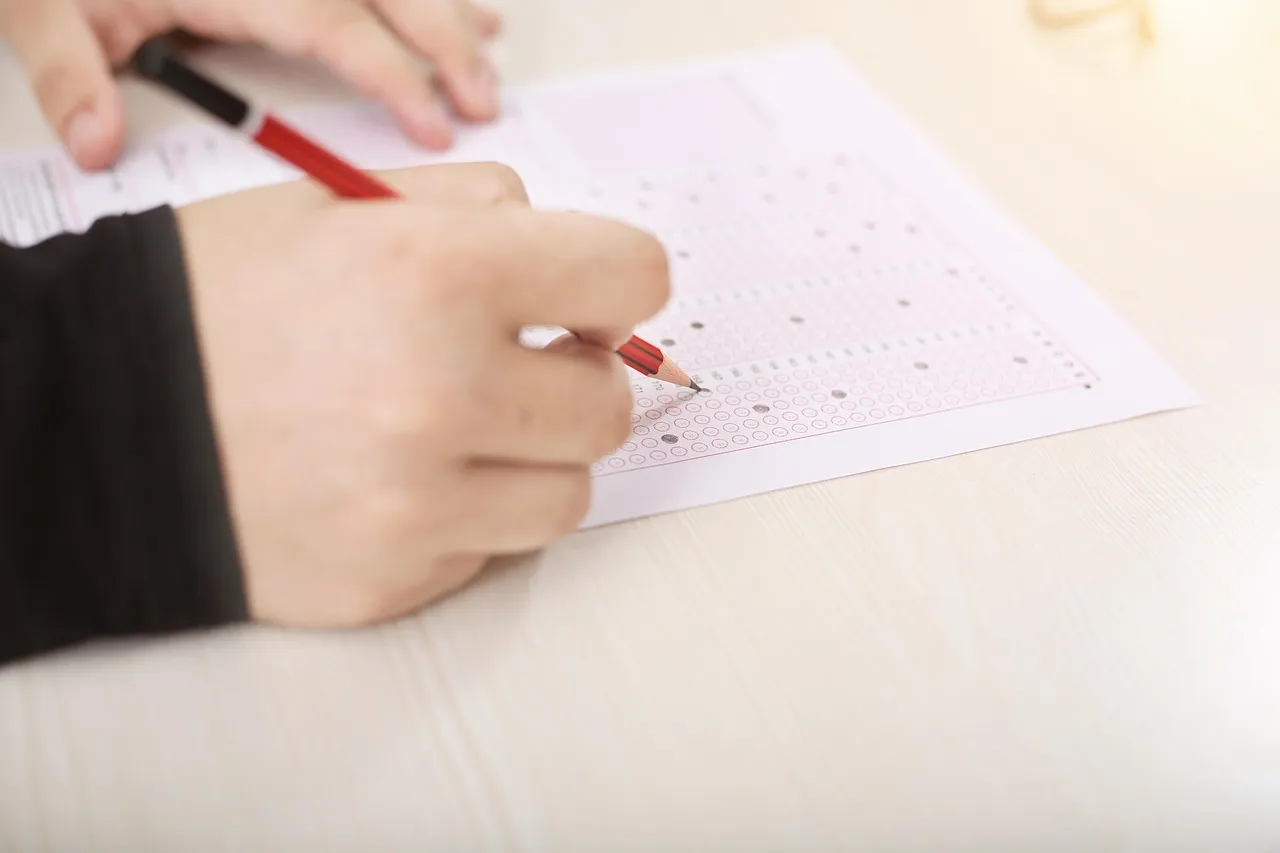 A hand holding a red pencil fills in one of the small oval bubbles on a printed multiple-choice answer sheet lying on a pale wooden desk.