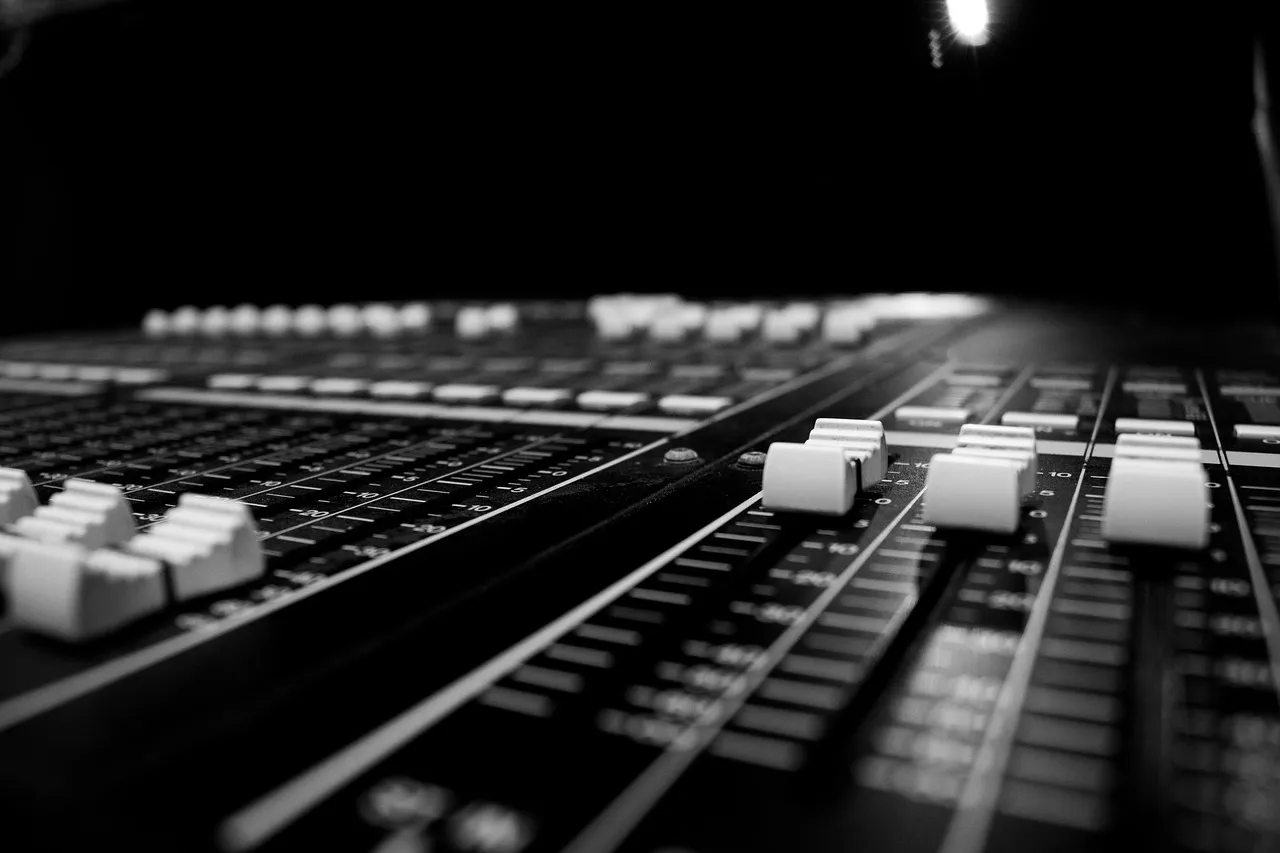A close-up of an audio mixing console, rows of faders receding into shadow