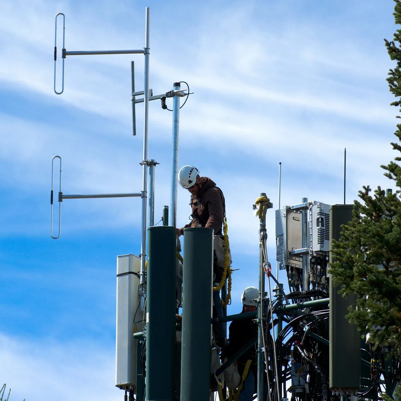 Two engineers in hard hats working among panel antennas at the top of a mobile phone mast against a blue sky