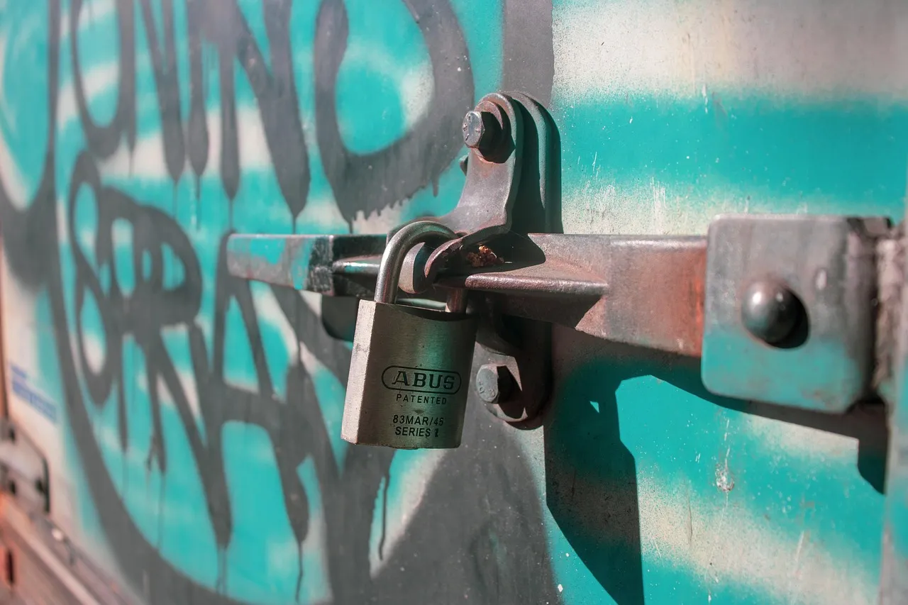 A plain brass padlock fastened through the steel latch of a shipping container door, photographed close up. The door behind it is painted teal and covered in graffiti, and the latch and bolt heads are scuffed with use.