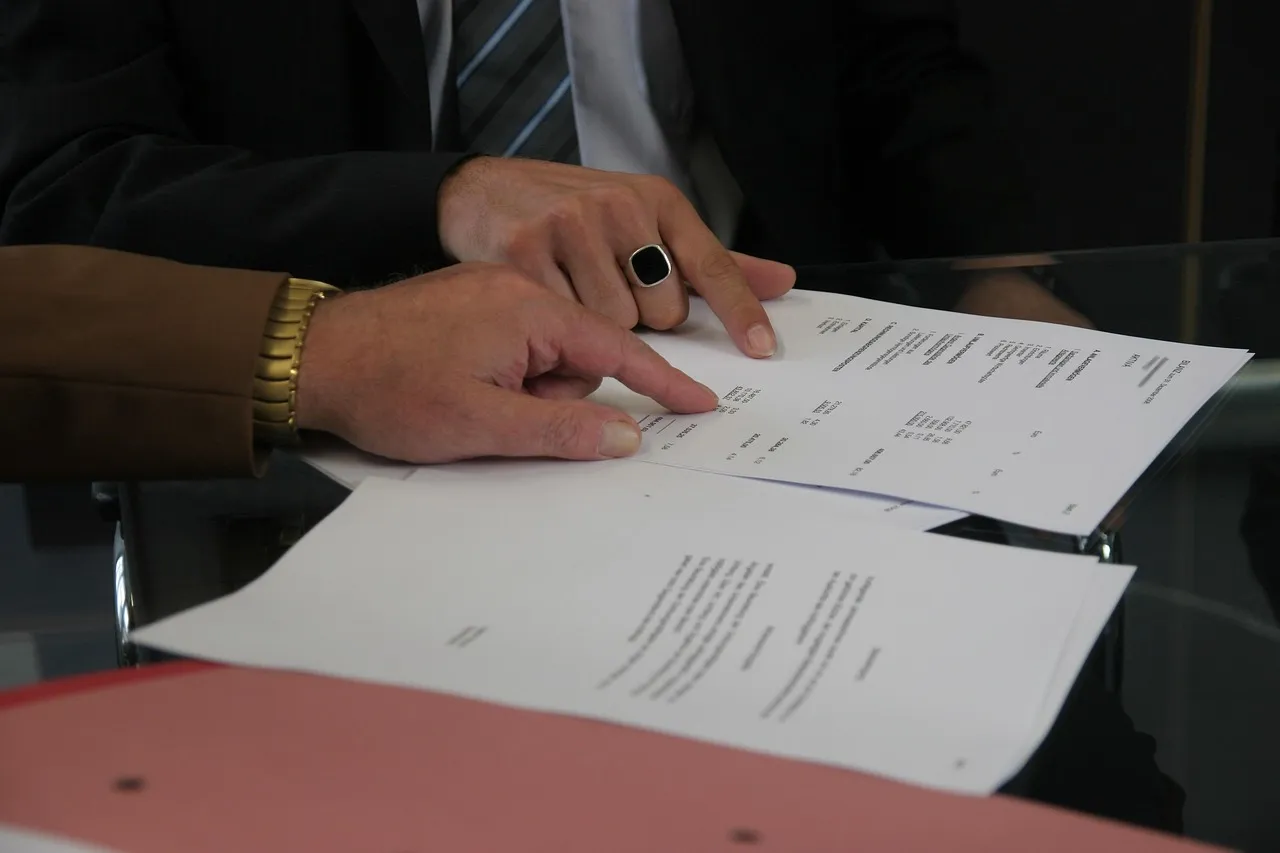 Two people at a glass table, one pointing at a specific line on a printed contract.