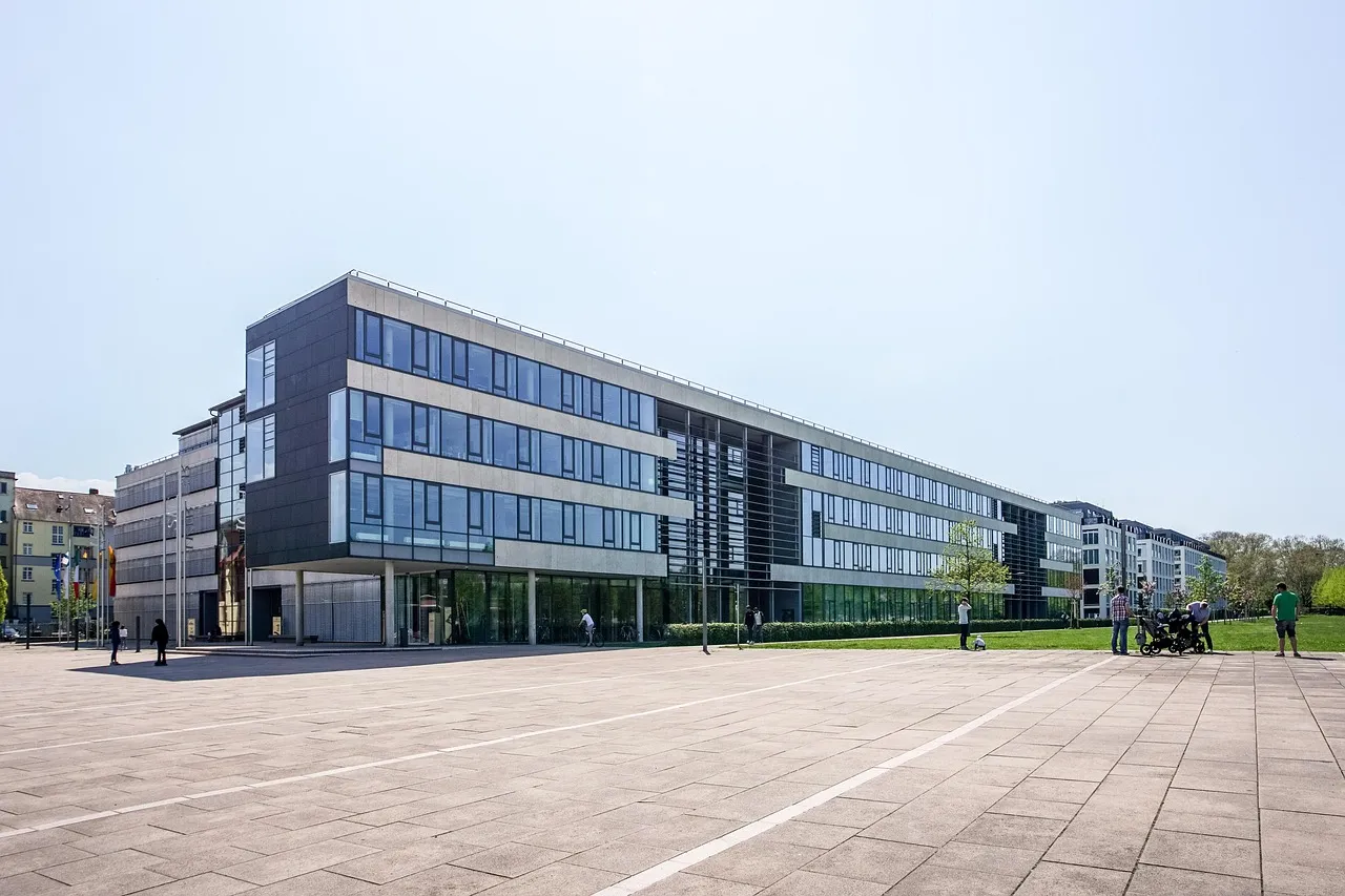 A modern multi-storey office building with long rows of windows, beside a wide paved plaza.