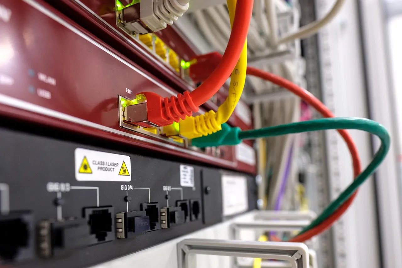 Red, yellow and green network patch cables plugged into the lit ports of a rack-mounted switch, with more cabling running out of focus behind it.