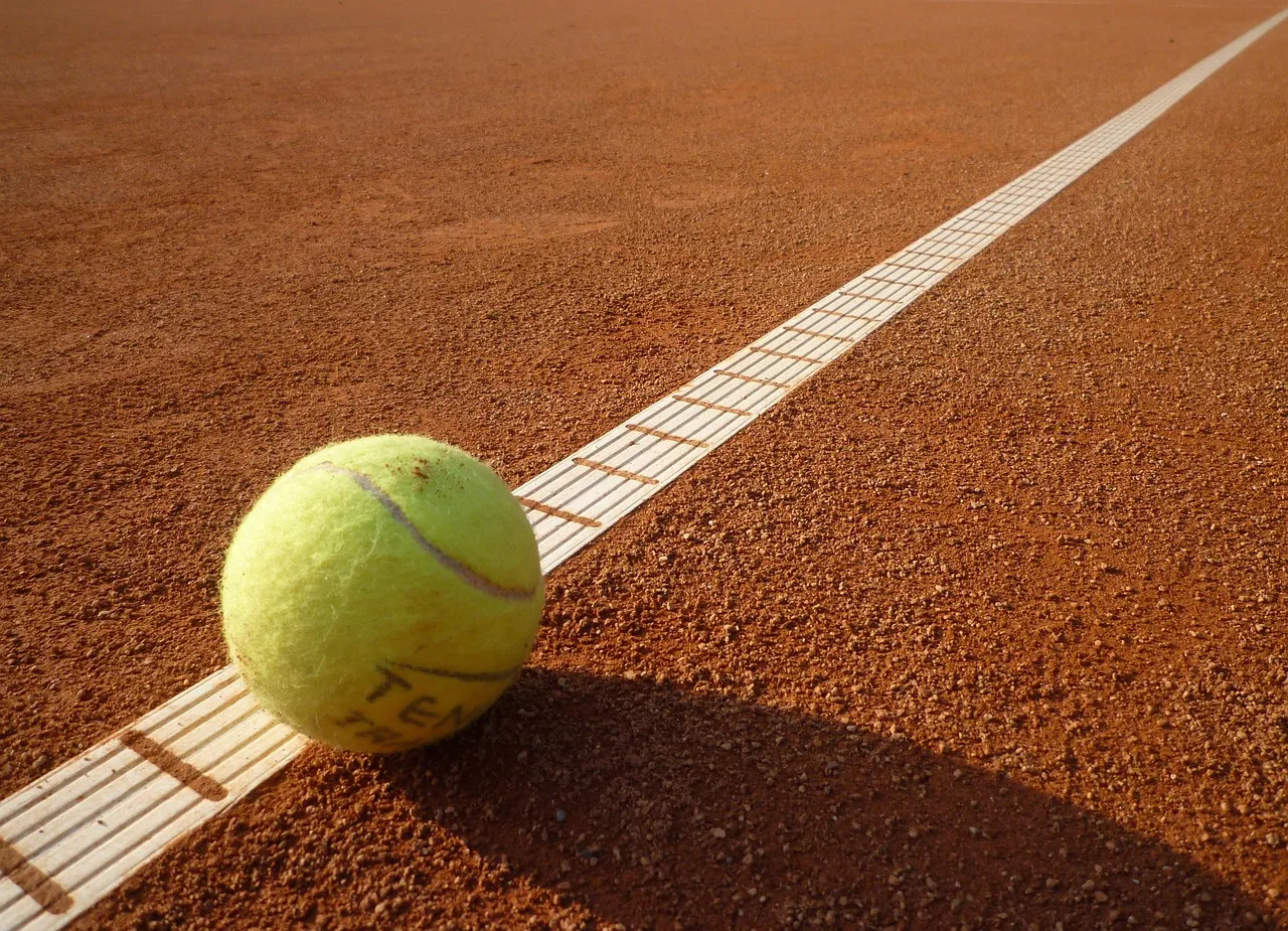 A tennis ball resting on the white boundary line of a clay court, half on each side of it.