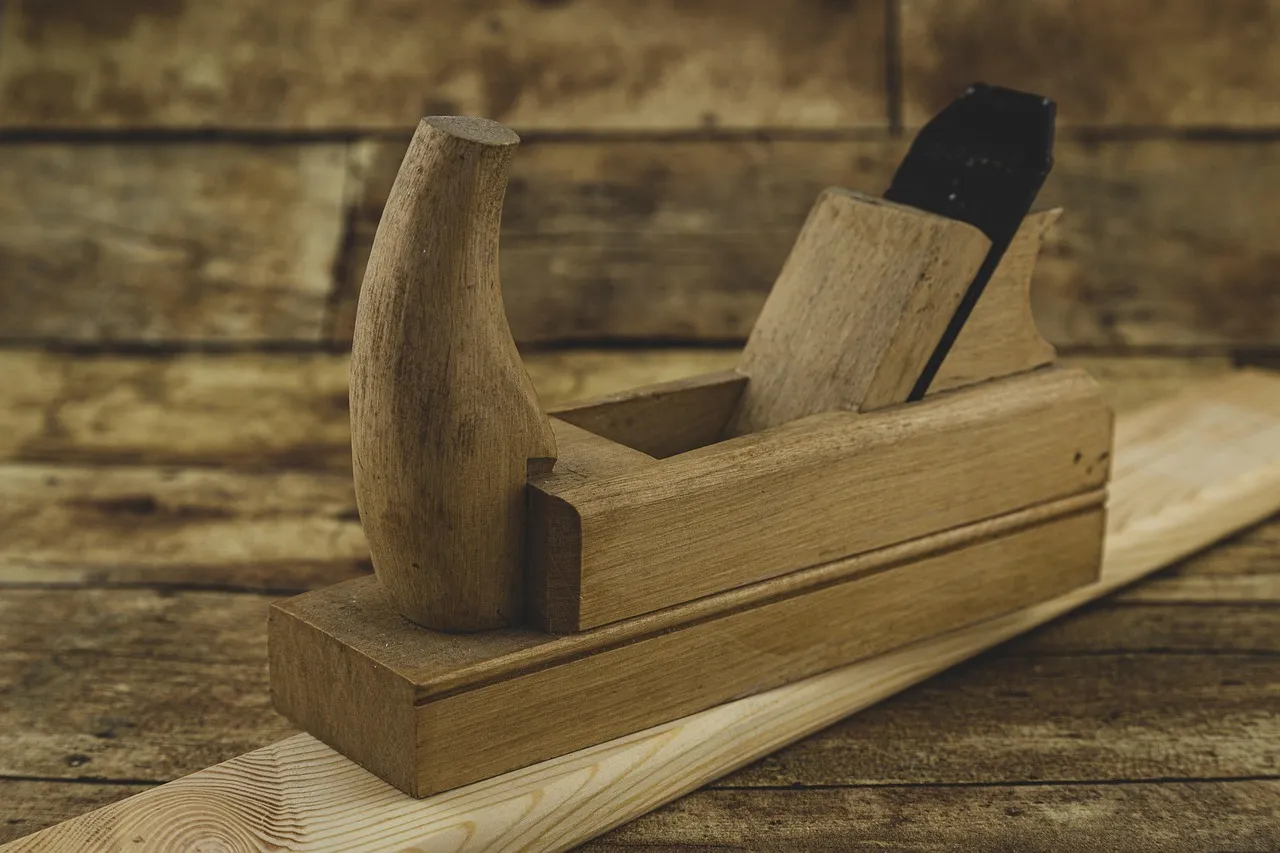 A wooden hand plane resting on a fresh length of planed timber against a weathered plank wall, its blade and wedge still seated and its handle worn smooth from use.