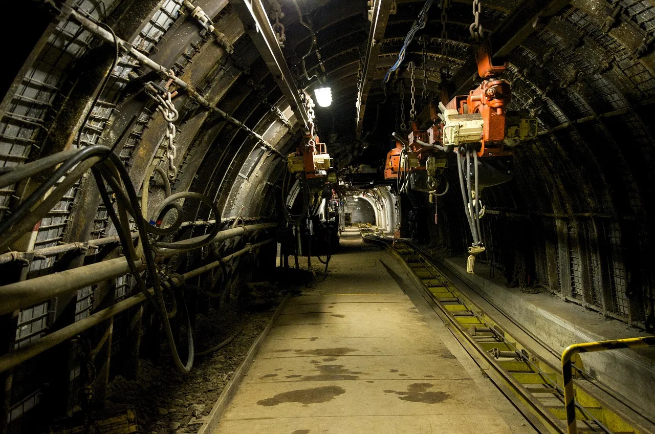 A lit tunnel deep underground, with rails running along the floor and pipes and machinery fixed along its arched roof.