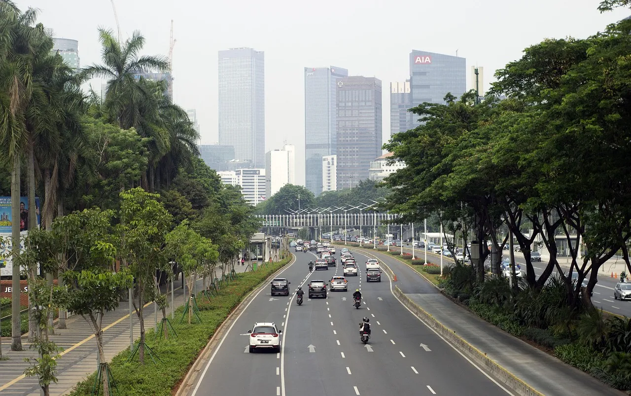 Traffic on a tree-lined Jakarta arterial road, office towers and a construction crane rising through haze behind it — illustrative of the Indonesian market CoreWeave is entering.