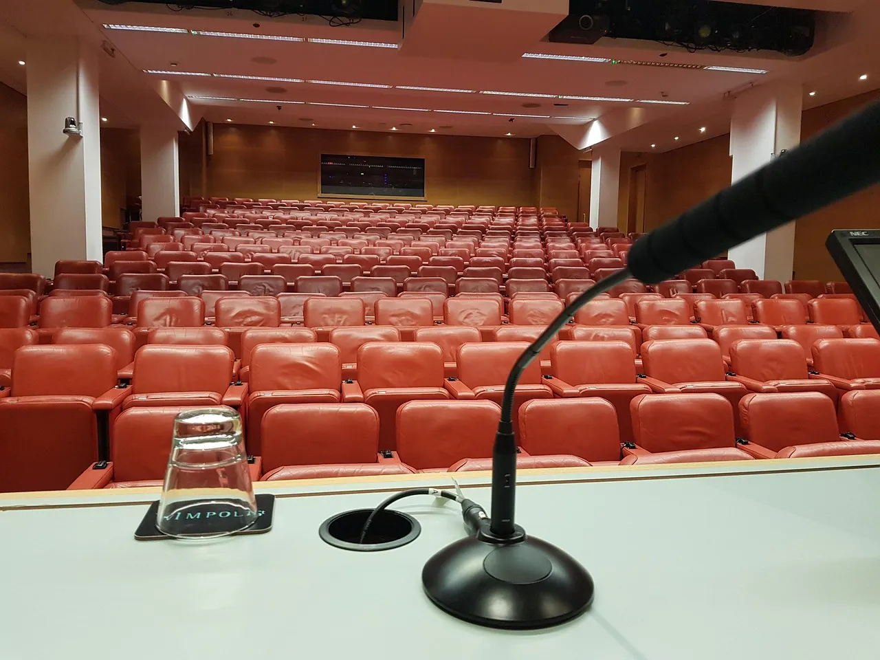 An empty conference auditorium of red seats seen from behind the speaker's lectern, with a gooseneck microphone and an upturned water glass in the foreground