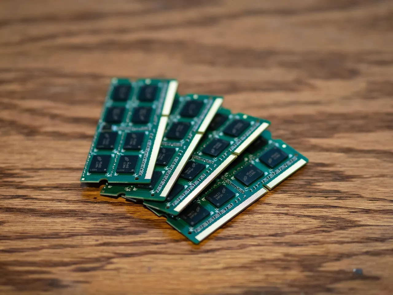 Four green memory modules fanned out on a wooden desk, their black chips and gold contact edges in close focus.