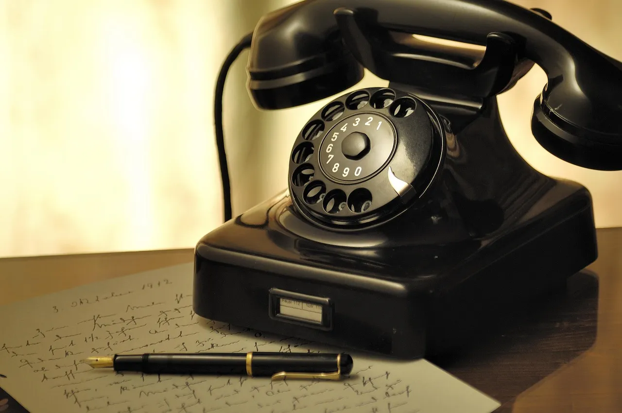 A black rotary telephone on a desk beside a fountain pen, lit from one side