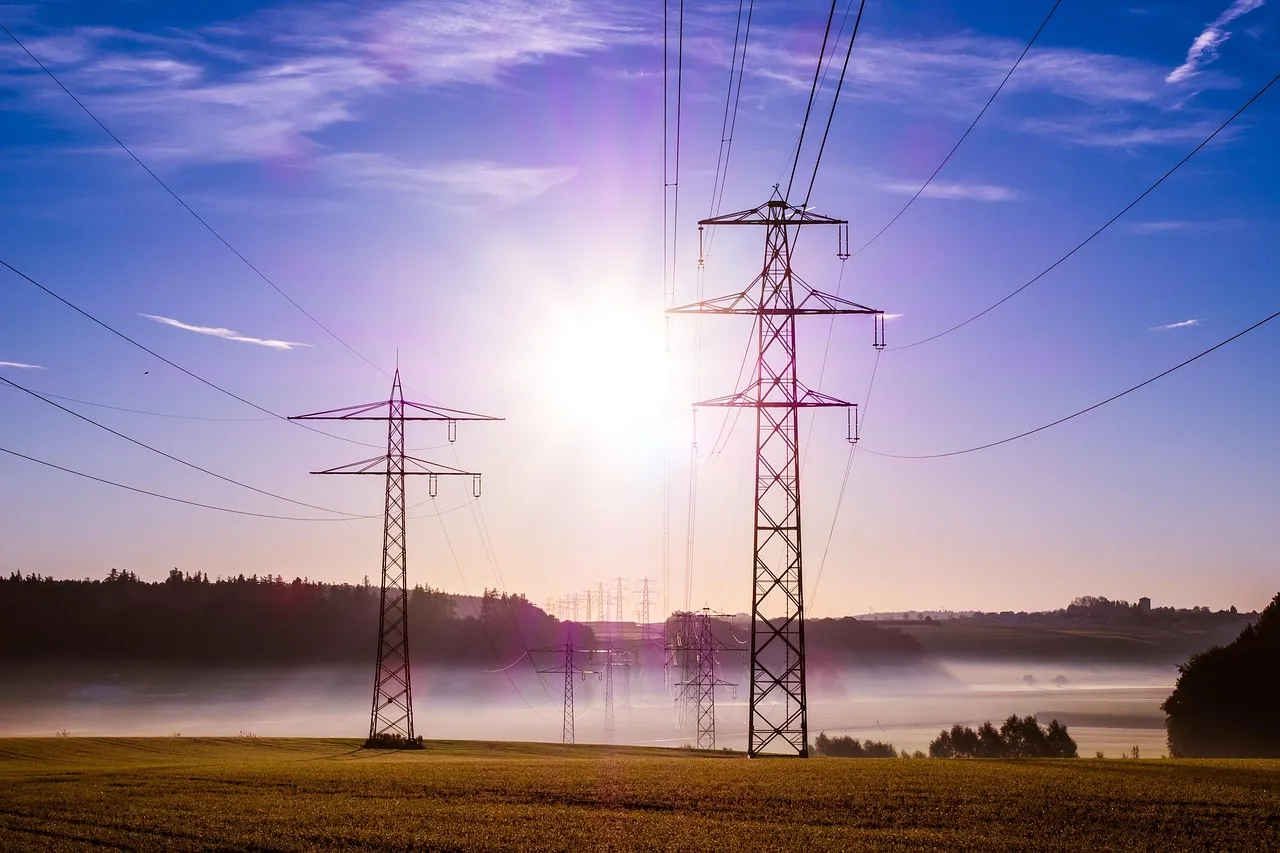 Electricity pylons and transmission lines across a misty field at sunrise