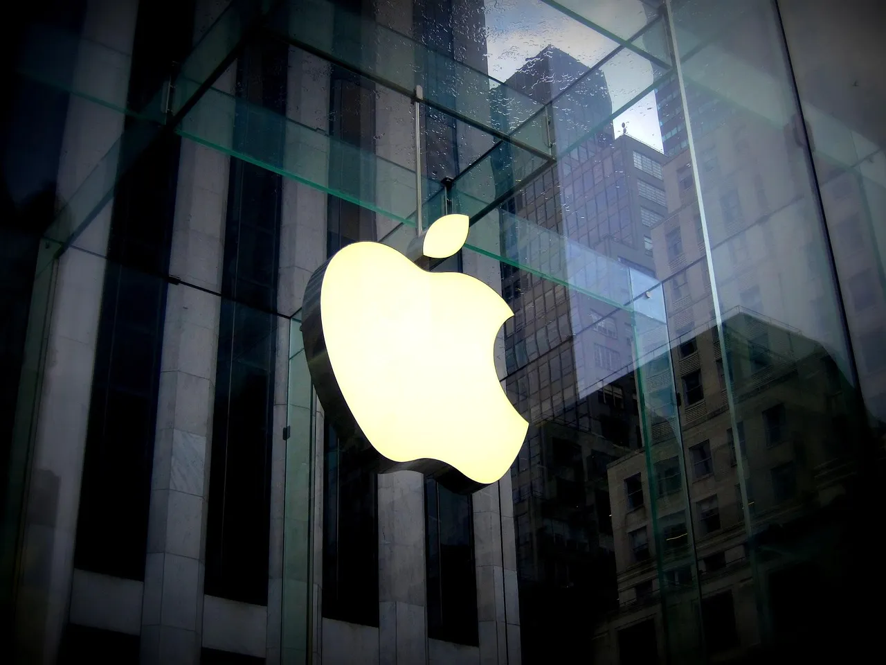 An illuminated Apple logo mounted on the glass facade of a store, with city buildings reflected in the panes