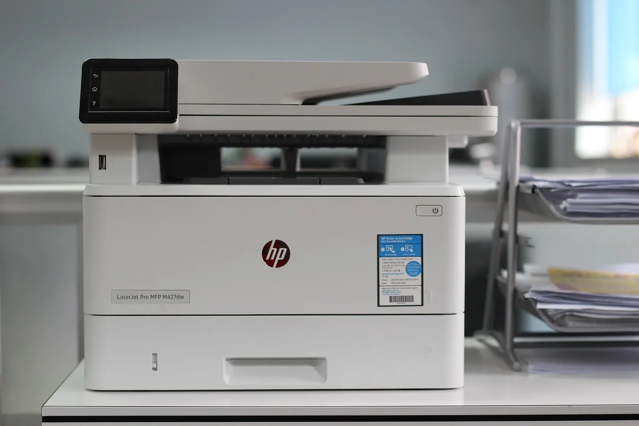 An office laser printer on a shelf beside stacked document trays