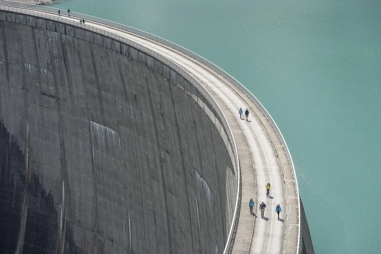 The curved concrete wall of a dam holding back a green reservoir, illustrating a report on water infrastructure funding for a data centre expansion.
