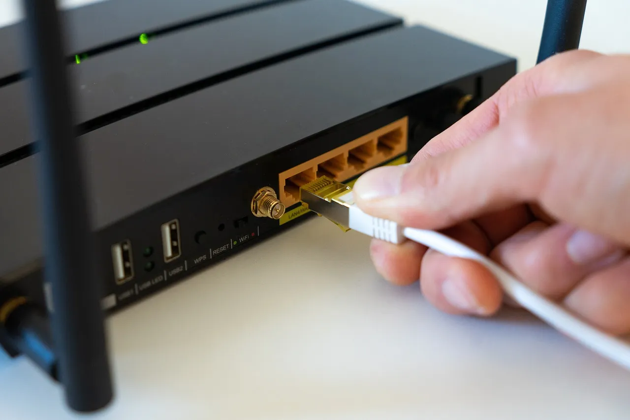 A hand plugging a white network cable into the LAN port of a black home router, illustrating a report on Macs exposed to the internet through remote access.