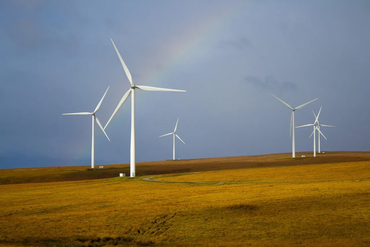 Wind turbines standing across open grassland under a dark sky and a faint rainbow