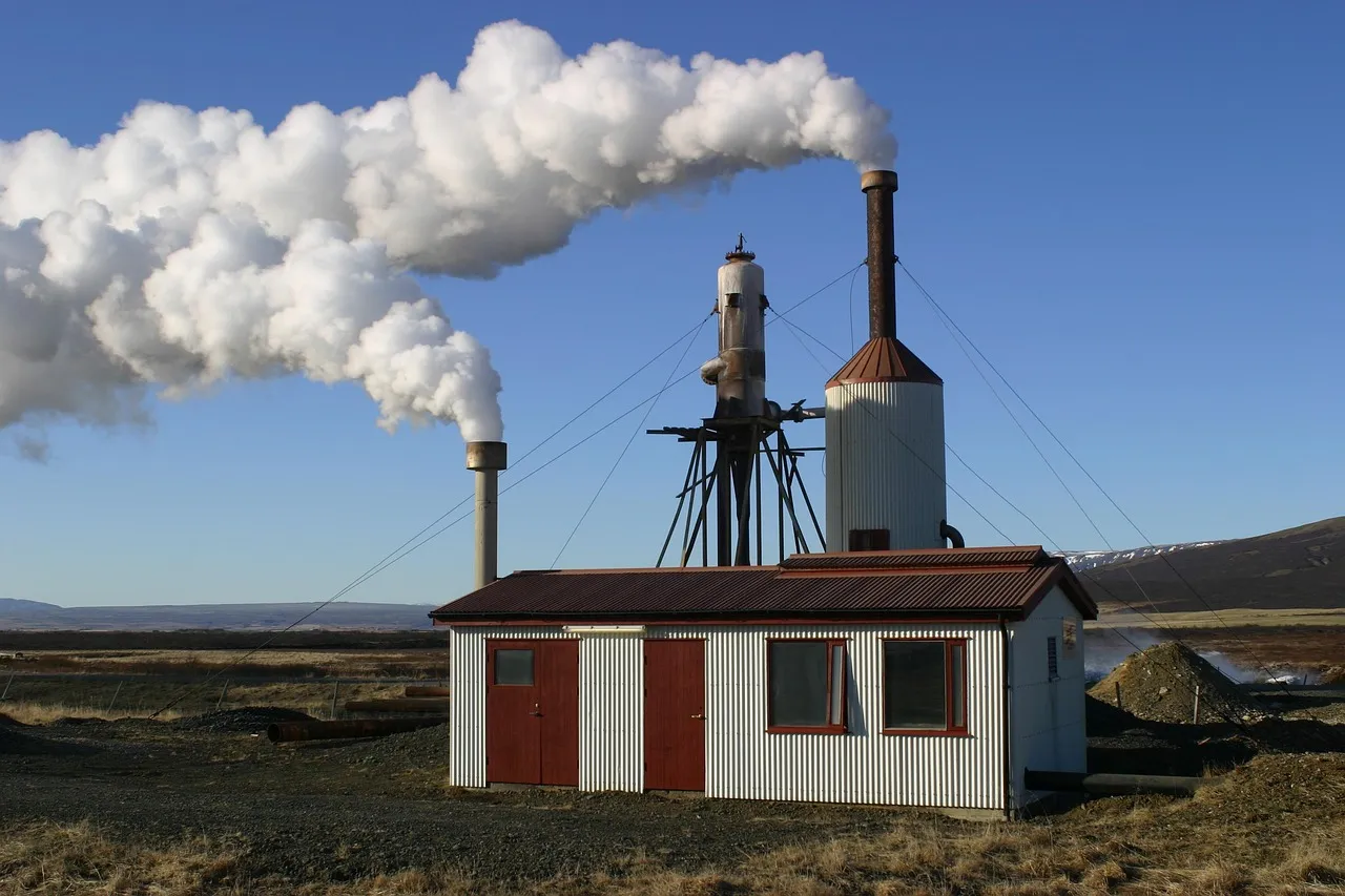 A small corrugated-metal geothermal wellhead building in open country, with a tall plume of steam venting from its stack