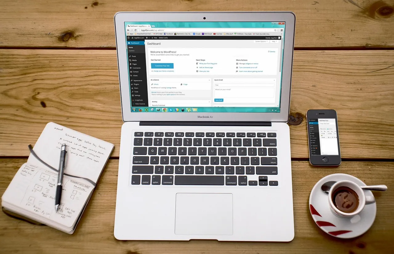 An overhead view of a laptop open on a wooden desk showing a web page, beside a notebook, a pen, a phone and a cup of coffee.