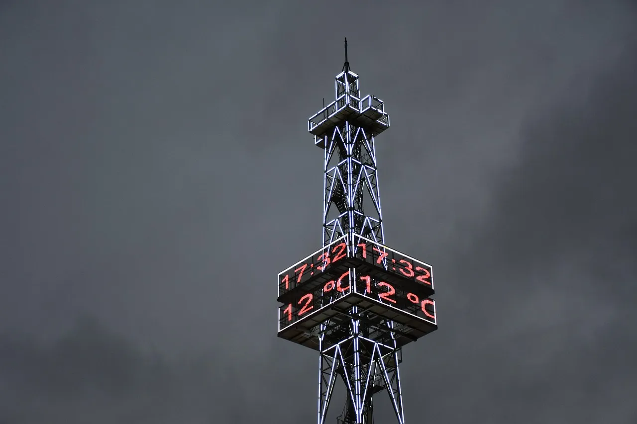 A lit steel telecommunications tower against a dark overcast sky, its display panel showing the time and temperature