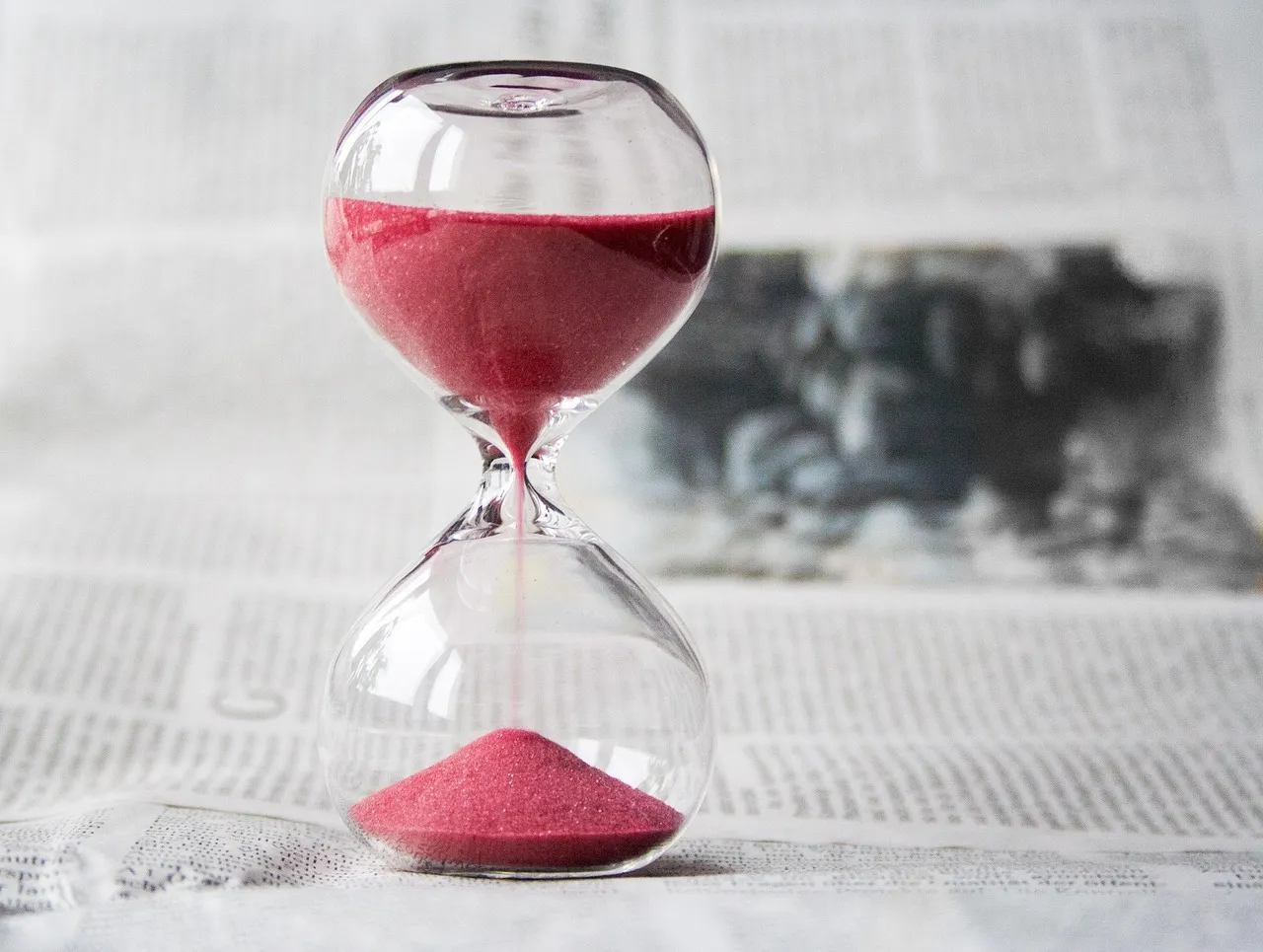 An hourglass with red sand running into the lower bulb, standing on a newspaper