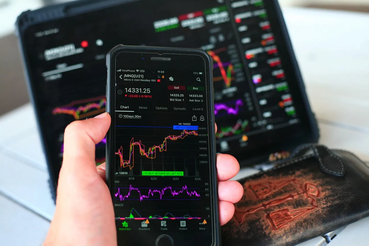 A phone showing candlestick price charts held in front of a monitor displaying more of the same.