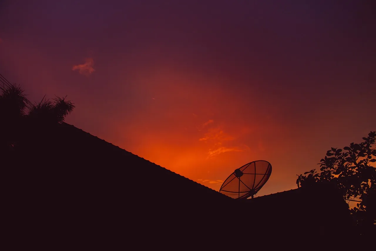 A satellite dish silhouetted on a rooftop against an orange sunset sky.